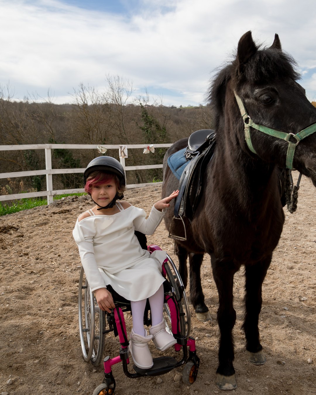 A young person in a wheelchair reaching up to stroke a pony in an outdoor arena